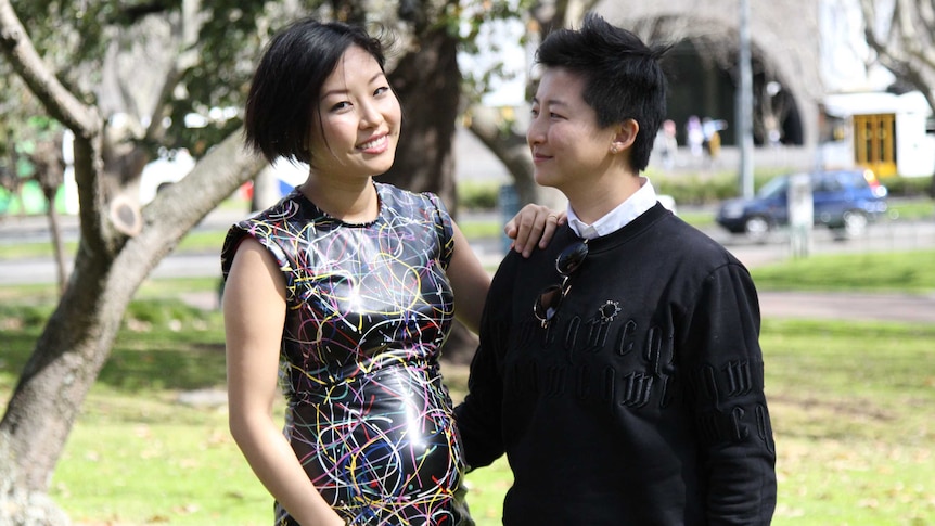 In a park, a pregnant Joyce looks at the camera while Tick looks at her.