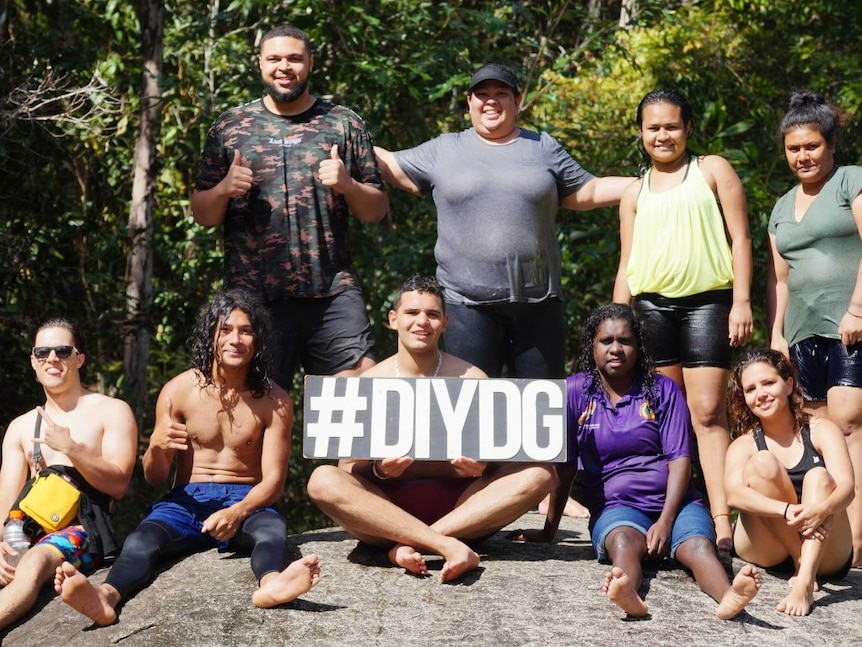 A group of nine people sit and stand on a rockface, with a man in the centre of those sitting holding a "#DIYDG" sign.