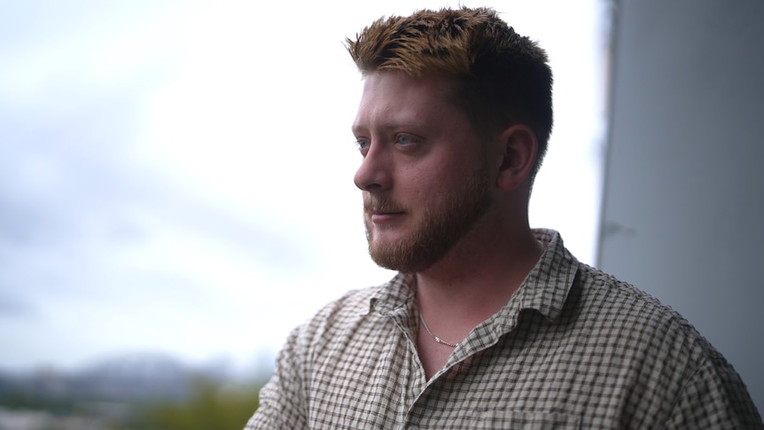 A young man with redish hair and wearing a checked shirt looks out from a balcony.