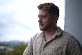 A young man with redish hair and wearing a checked shirt looks out from a balcony.