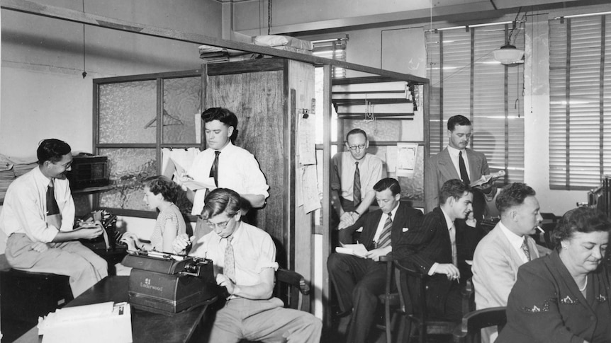 Black and white photo of newsroom in 1960s, group of people sit at typewriters working