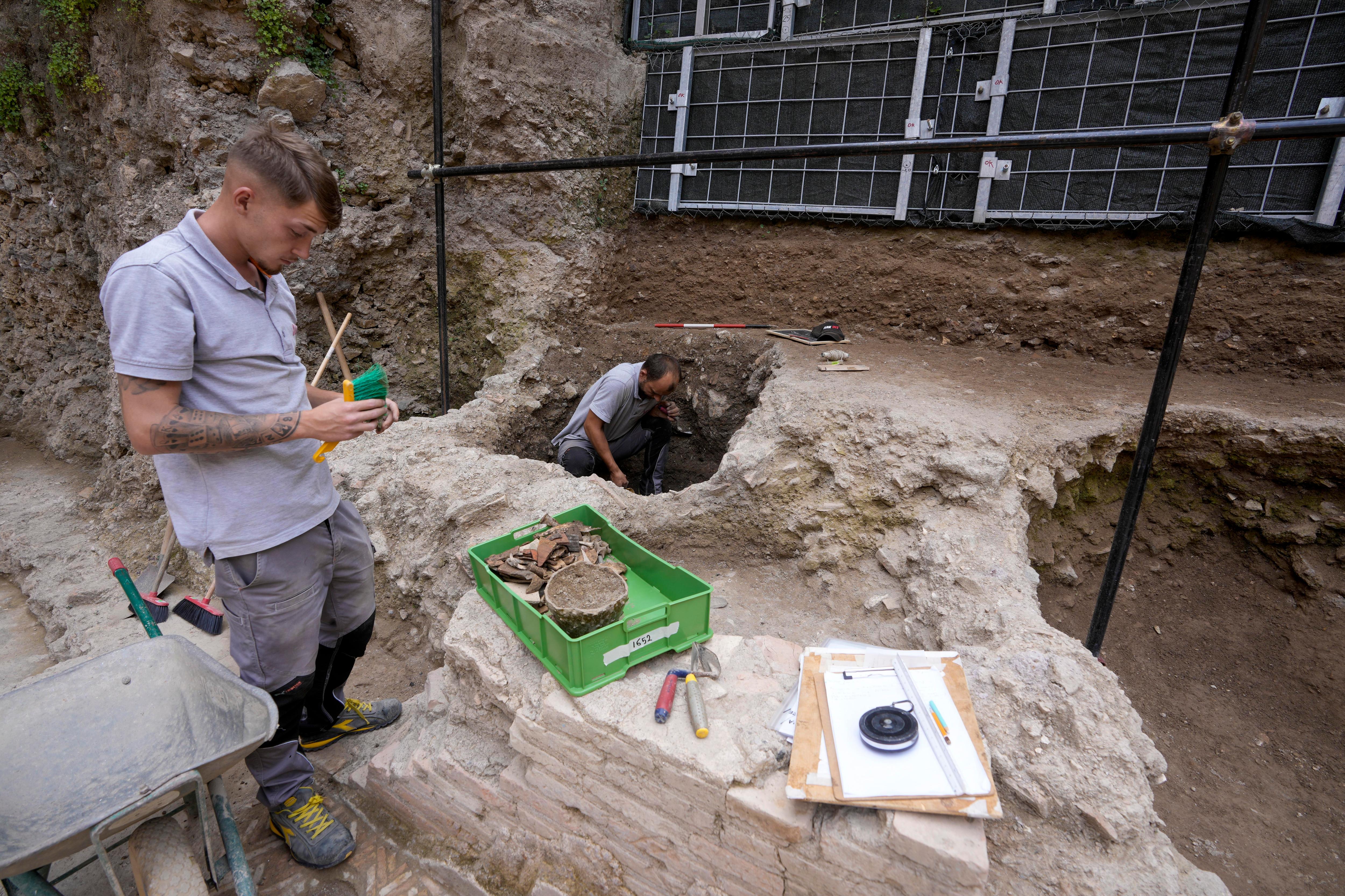 A man standing in an archaeological dig site holding a brush with other tools and a man around