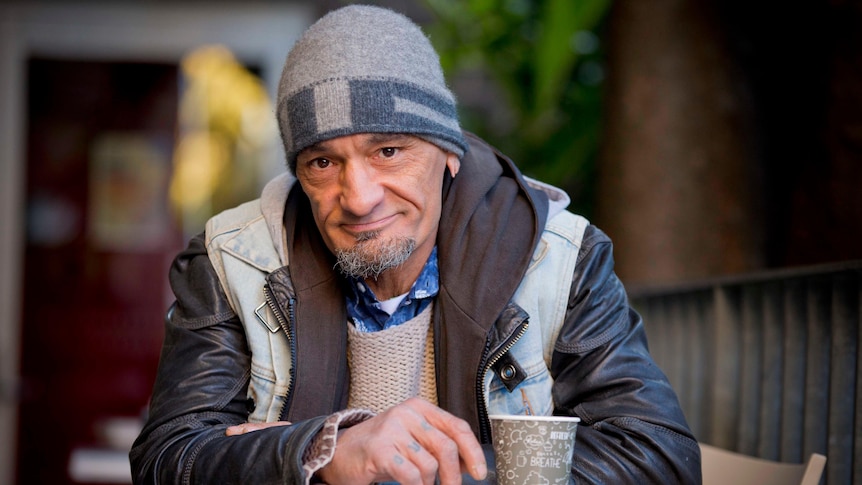 A man with a beanie and jacket sitting at an outdoor table, looking at the camera