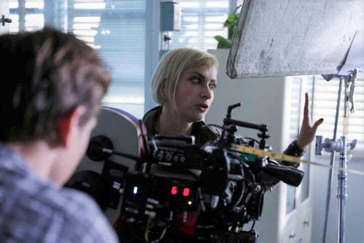 A woman with short blonde hair on the set of a movie surrounded by camera equipment 