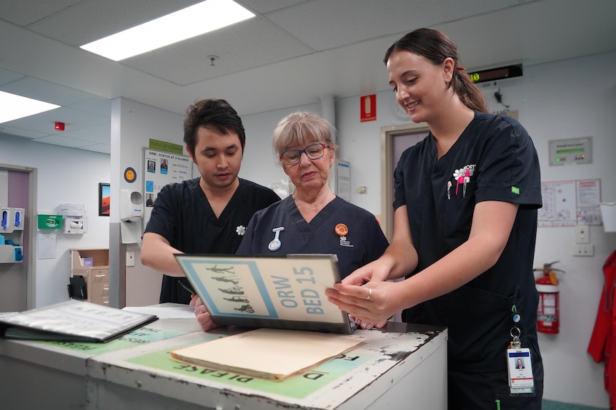 An older woman stands in the middle of two nurses in their 20s looking at patient information.