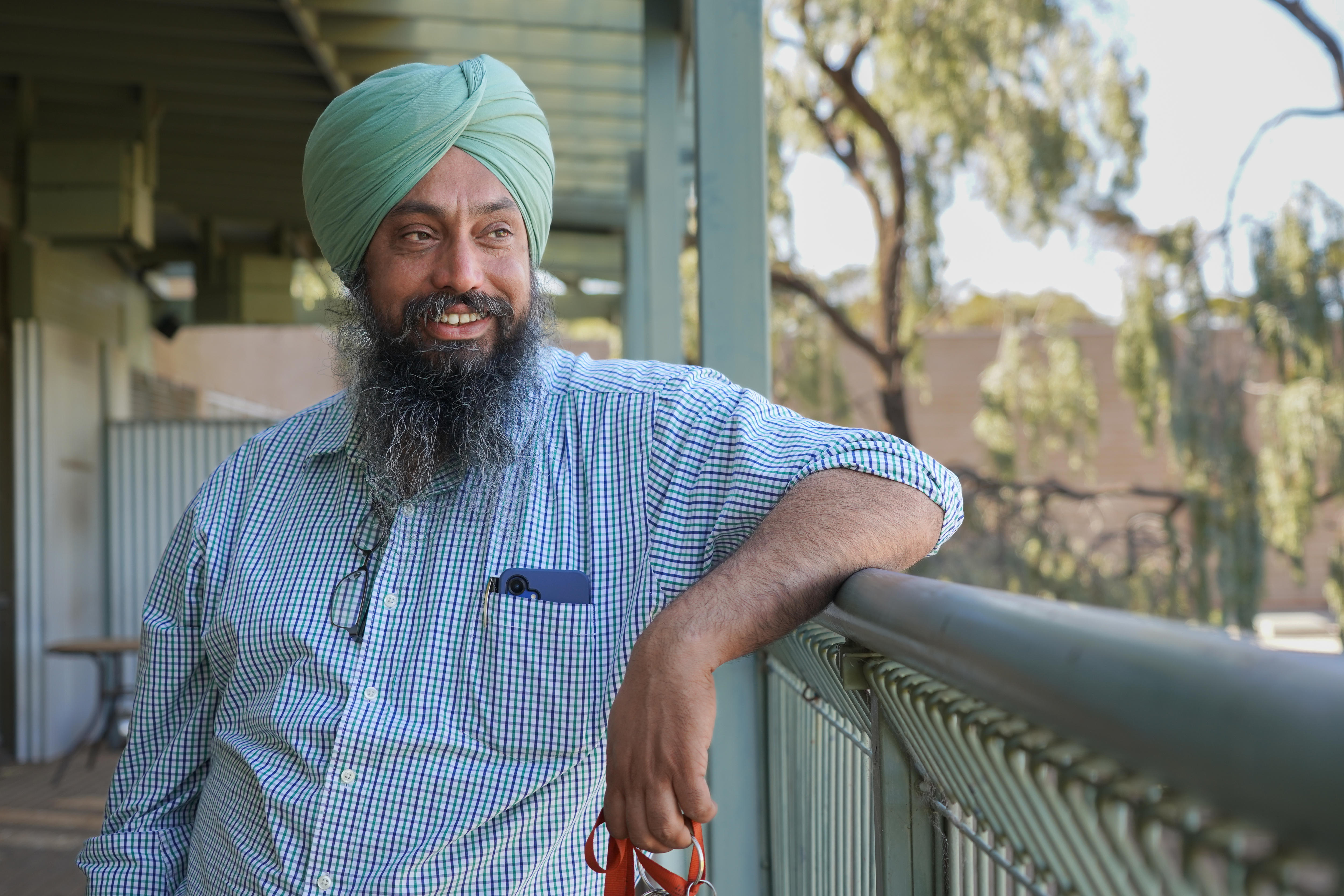 A man in a checked shirt and turban stands outside with his elbow leaning on a balcony railing starting off into the trees.