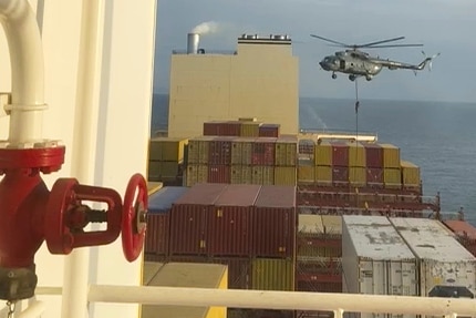 A helicopter hovers above container ships with a soldier moving down a rope toward them