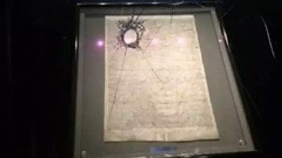 Blurry image of a Magna Carta behind broken glass in a dark room.