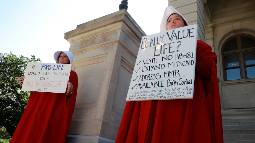 Two women hold protest signs while dressed as Handmaids in red dresses and white hats.