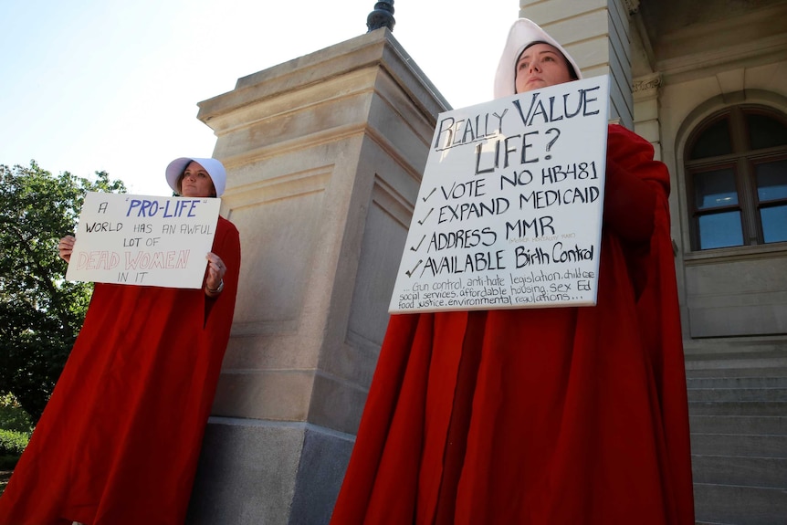 Two women hold protest signs while dressed as Handmaids in red dresses and white hats.