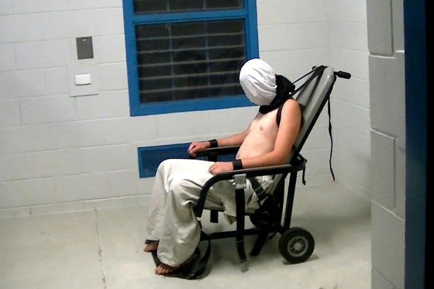 A topless young man is strapped to a chair with a hood over his head.