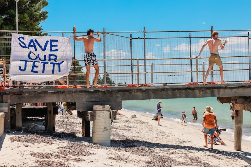 Two teen boys in boardshorts climbing along jetty, hanging onto fence, 'Save our Jetty' sign on fence