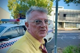 A middle-aged man with grey hair and glasses standing near a police car.