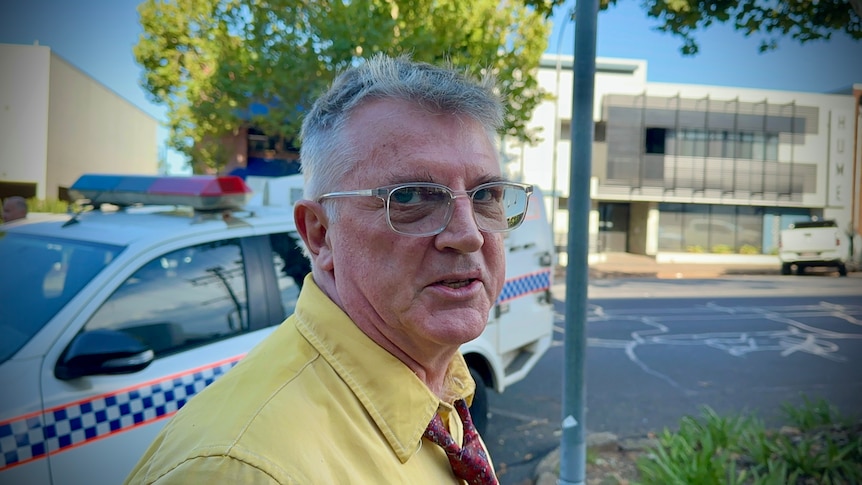 A middle-aged man with grey hair and glasses standing near a police car.