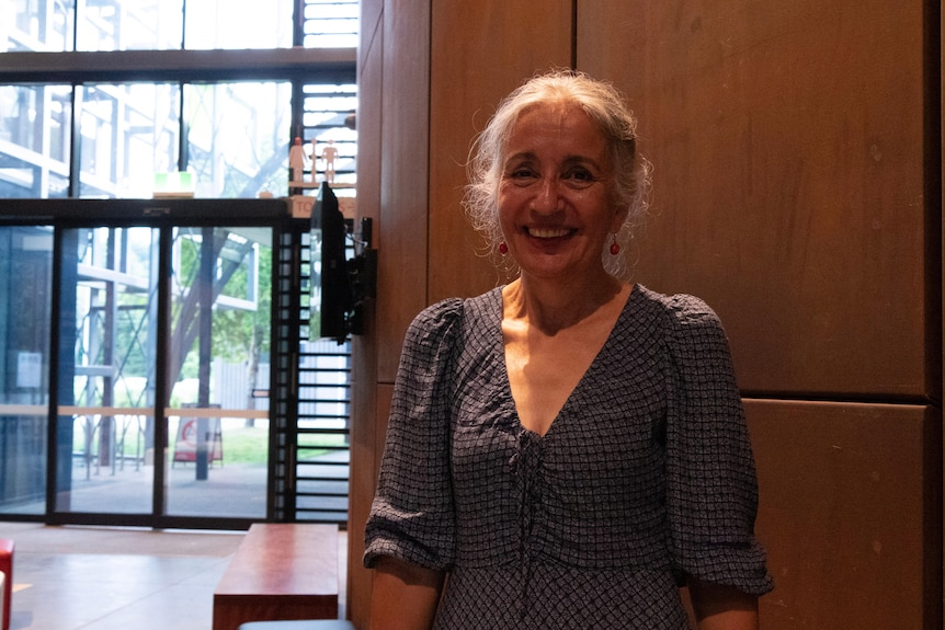 A woman with grey hair and a blue dress stands smiling in front of a wall