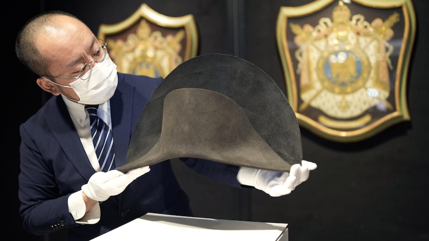 An auction house employee in white gloves holds up the biocorne winter hat above its display case. 