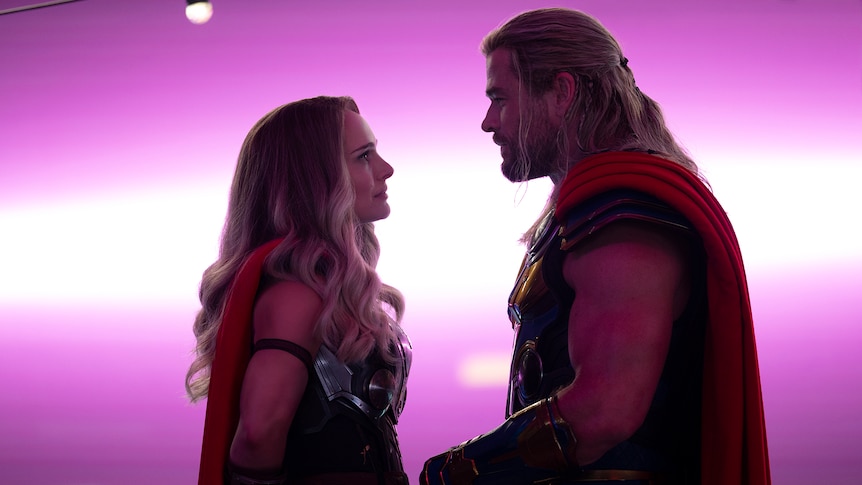 Blonde white woman stands looking up at blonde white man in neon pink lit space. Both wear armor and capes.