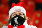 A woman wearing a Liverpool FC face mask and santa hat that says Ho! Ho! Ho! grimaces as she watches football