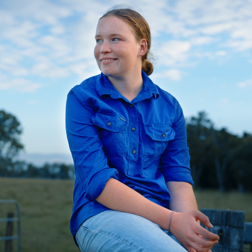 young girl sits on a cattle fence wearing a blue button down shirt and jeans. She looks away from the camera into the distance