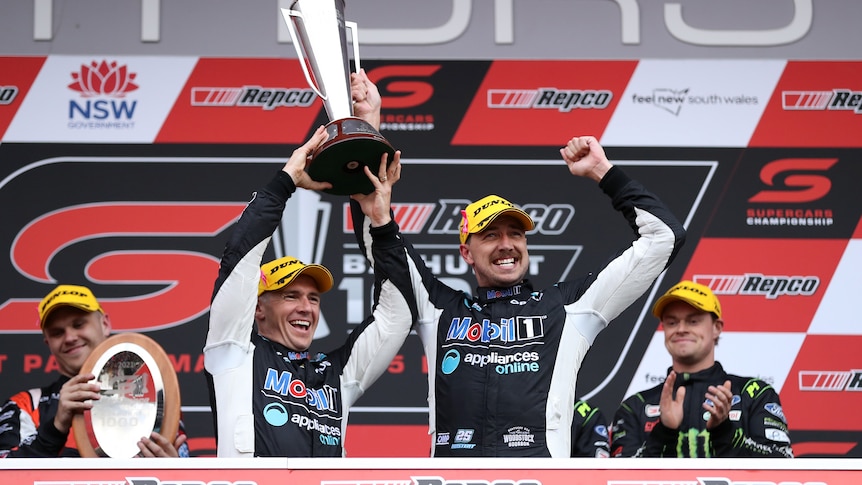 Two Supercars drivers stand on the podium smiling, holding the Bathurst 1000 trophy with one of them punching the air.