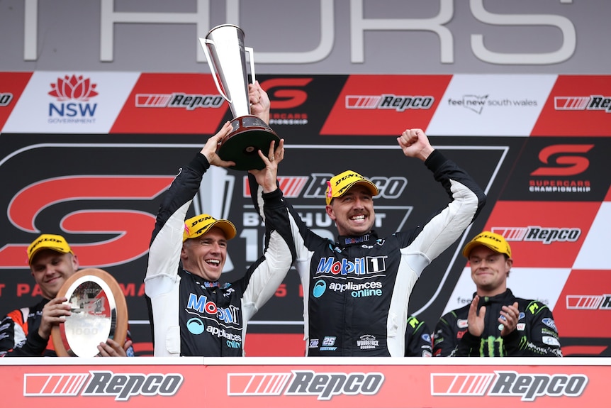 Two Supercars drivers stand on the podium smiling, holding the Bathurst 1000 trophy with one of them punching the air.