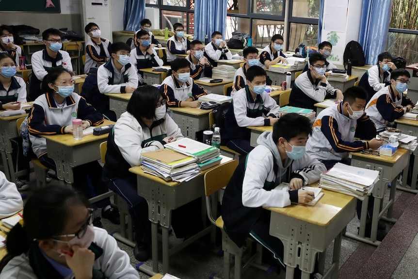 A woman wearing a face mask stands at a chalk board in front of students at desks in a classroom.