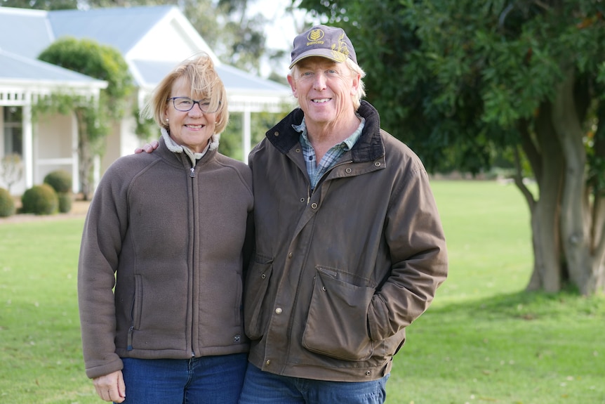 A smiling man and woman standing together in front of a beautiful farm house surrounded by green grass