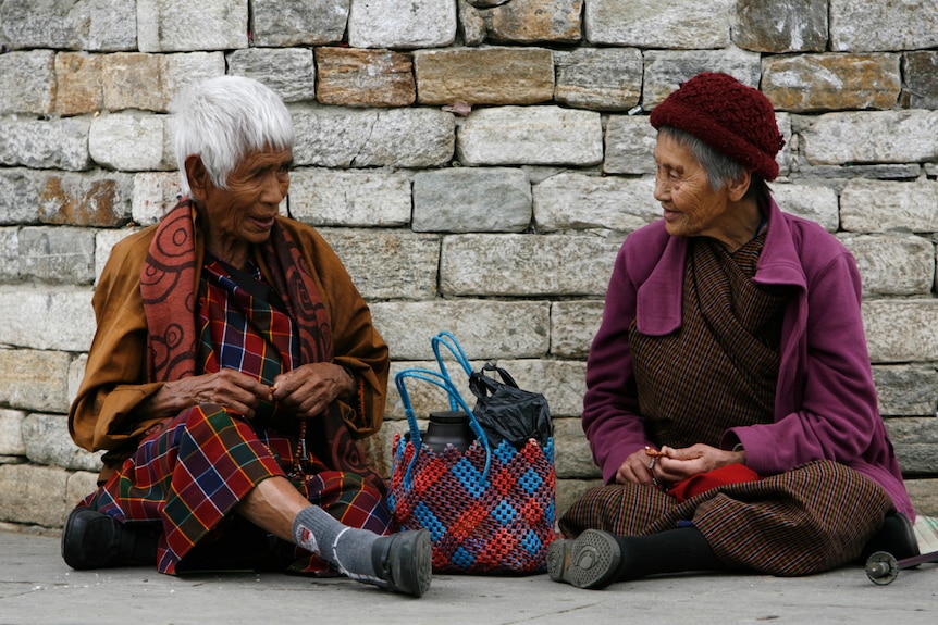Two old Bhutanese women sit next to each other on footpath.