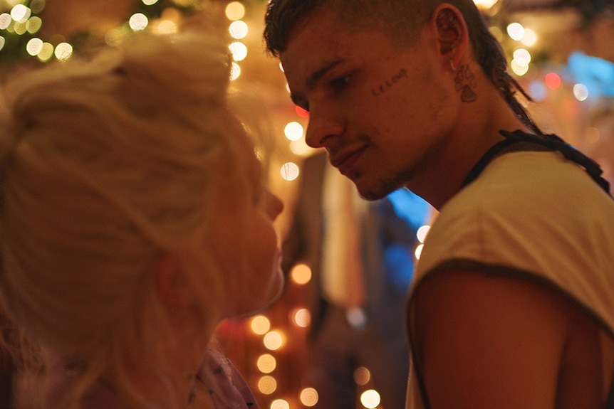 A young blonde girl and tattooed boy with rat tail stand close in party setting, behind them is out of focus fairy lights.