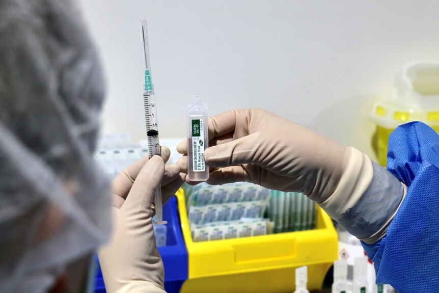 A close-up shot of someone's PPE-clad hands holding a syringe and a dose of the Pfizer COVID-19 vaccine.