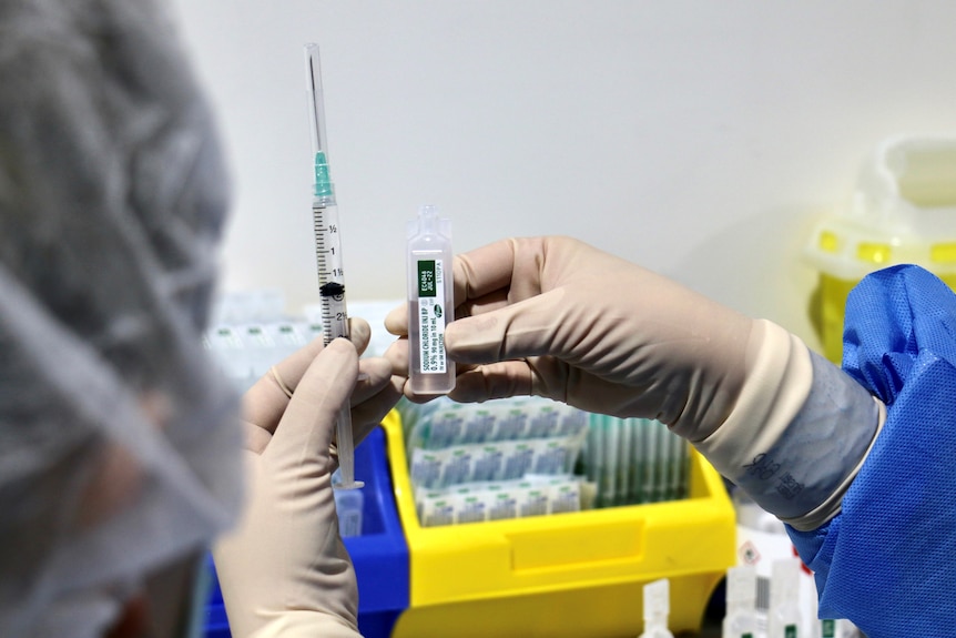 A close-up shot of someone's PPE-clad hands holding a syringe and a dose of the Pfizer COVID-19 vaccine.