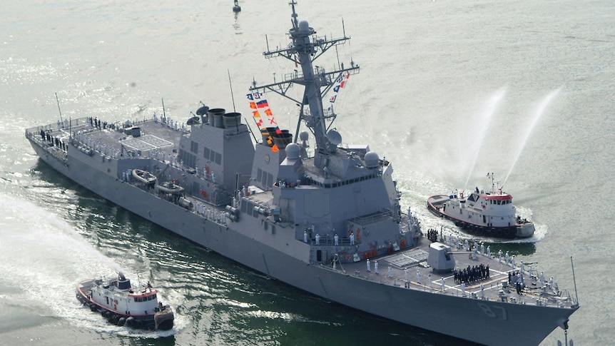 A warship with crew across its deck sails between two smaller vessels