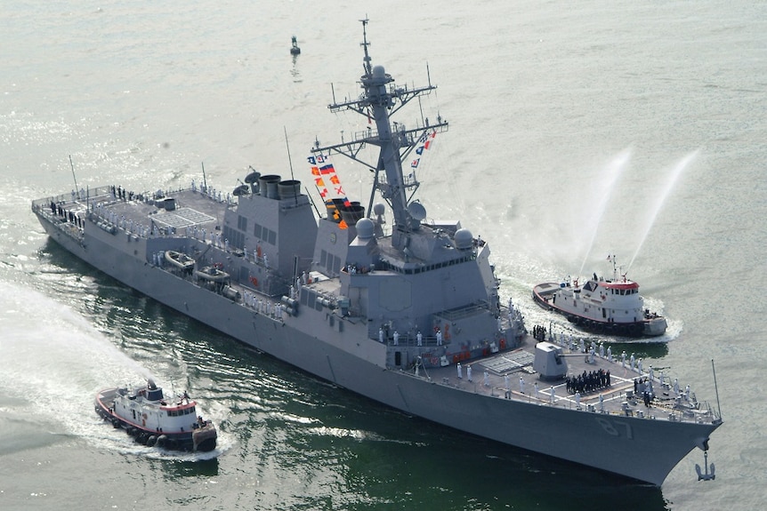 A warship with crew across its deck sails between two smaller vessels