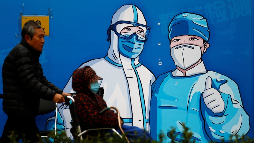 A man pushes a woman in wheelchair past a poster with a graphic of medical workers.