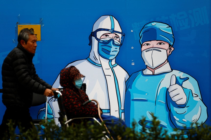 A man pushes a woman in wheelchair past a poster with a graphic of medical workers.