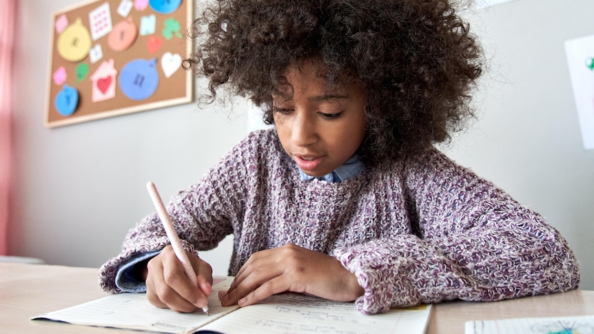 A young Black girl sits at a desk and writes in an exercise book.