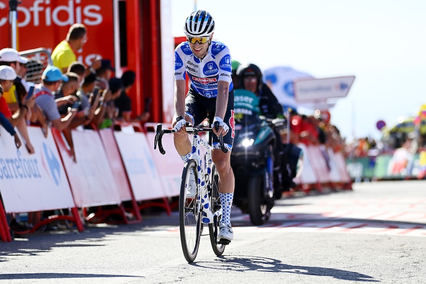 A smiling Australian cyclist in a blue and white polkas dot jersey rolls over the finish line on a stage of the Vuelta. 