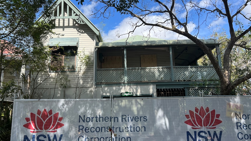 weatherboard two storey home with mesh security fence around it with Northern Rivers Reconstruction Corporation signage 