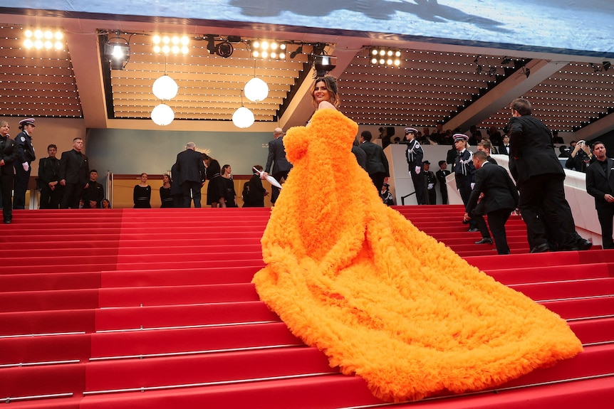 Deepti Sadhwani wearing a bright yellow-orange puffy coat with a long train, draping down a set of stairs