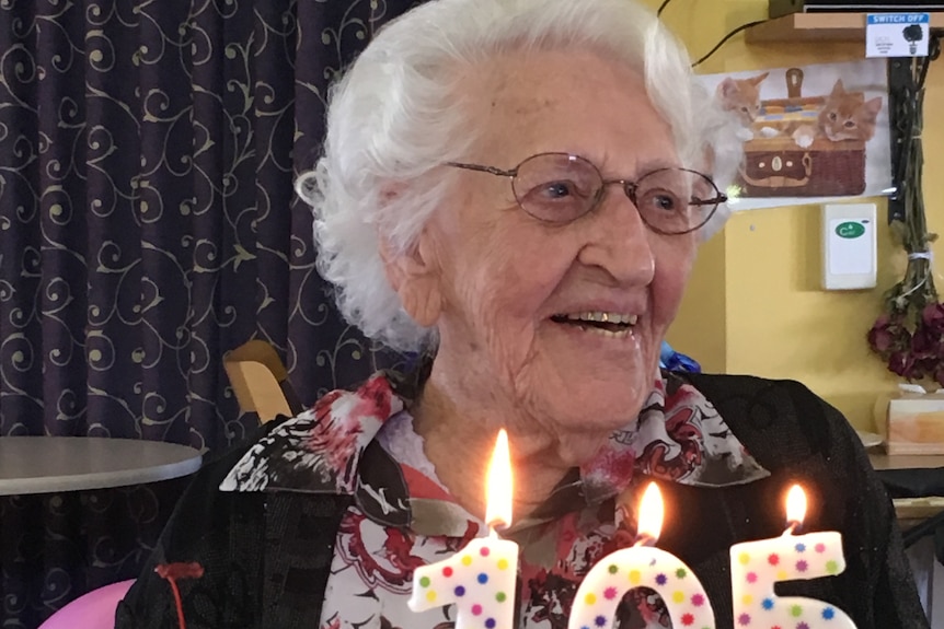 An older woman smiles as she sits behind a cake cake showing numerals 105.