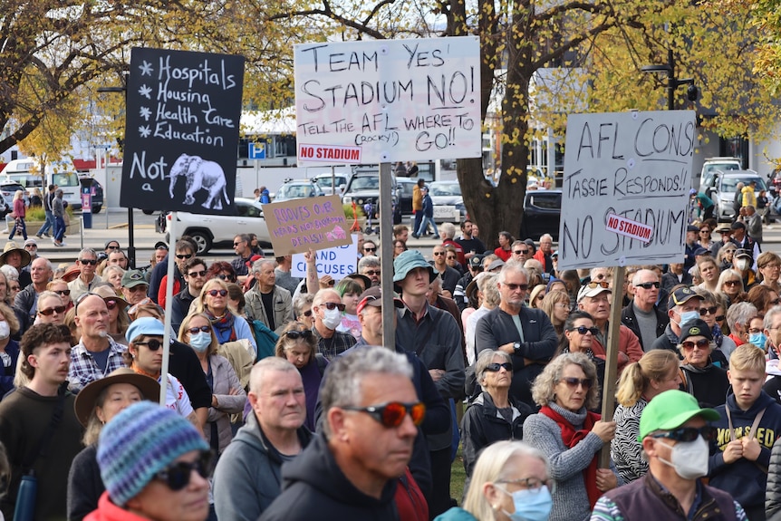 A crowd of people hold signs at a rally saying 'no stadium'