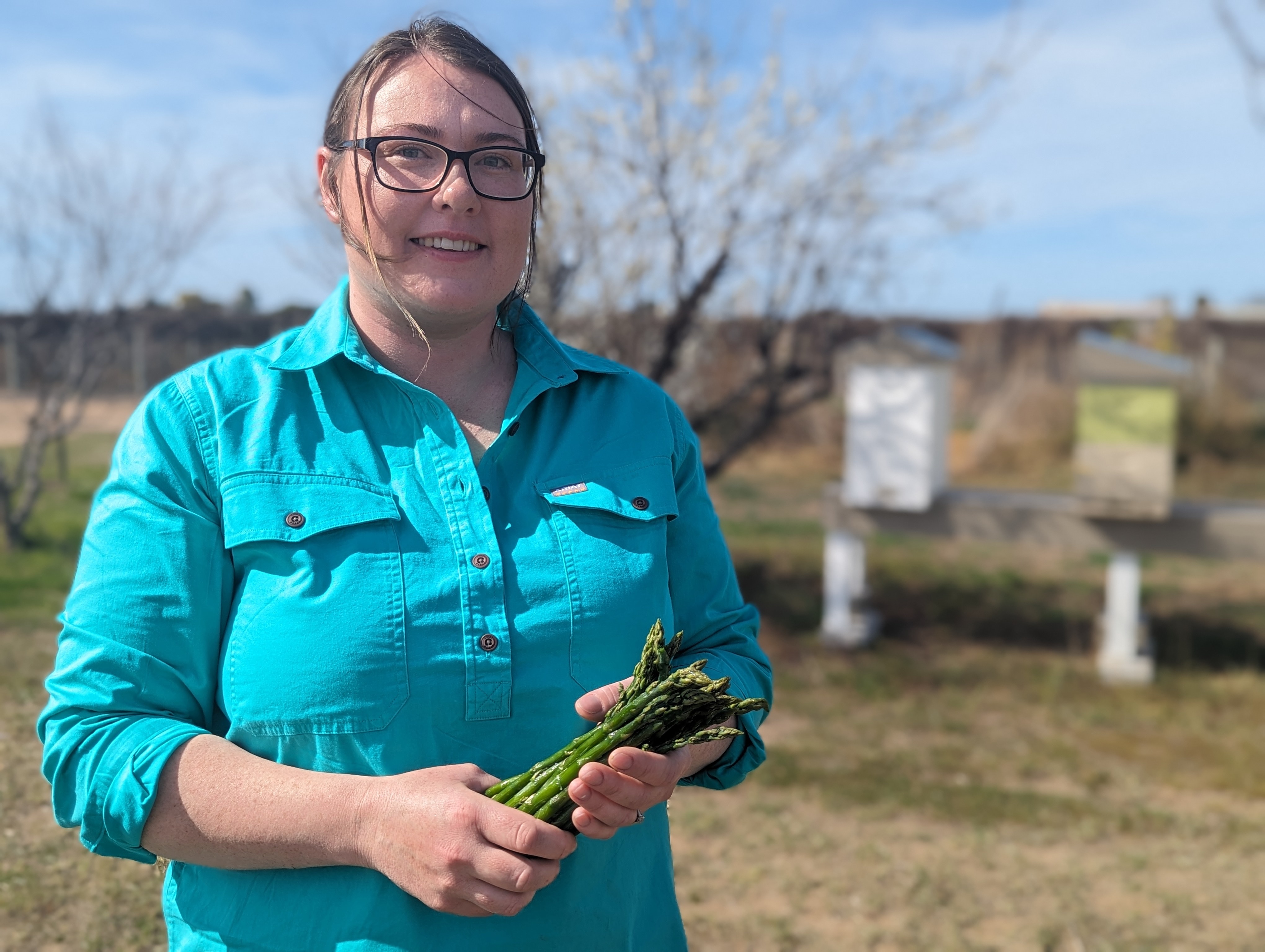 A fair-skinned woman, Renee, in black glasses and turquoise shirt holds a bunch of green asparagus in front of bee hives.