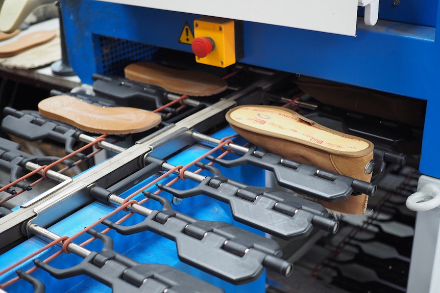 An upside down sheepskin boot and two leather soles go along a conveyor belt into a blue machine.