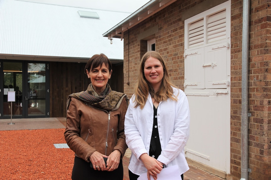 Megan Monte and Julie Gillick smile while standing outside the gallery with a brick wall behind them.