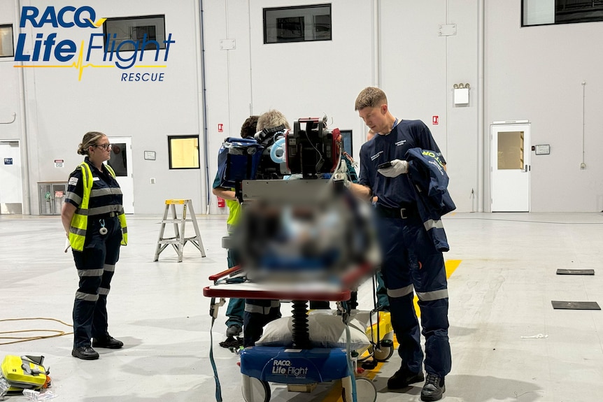 Paramedics stand around a bed in a hangar-like space.