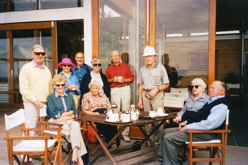 A group of older men and women wearing hats and sunglasses smile for a picture on an outdoor deck.
