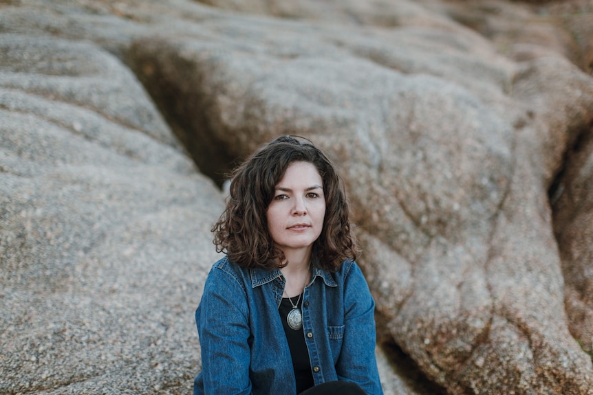 A press shot of Eliza Hull in a denim jacket sat amongst smooth rock formations