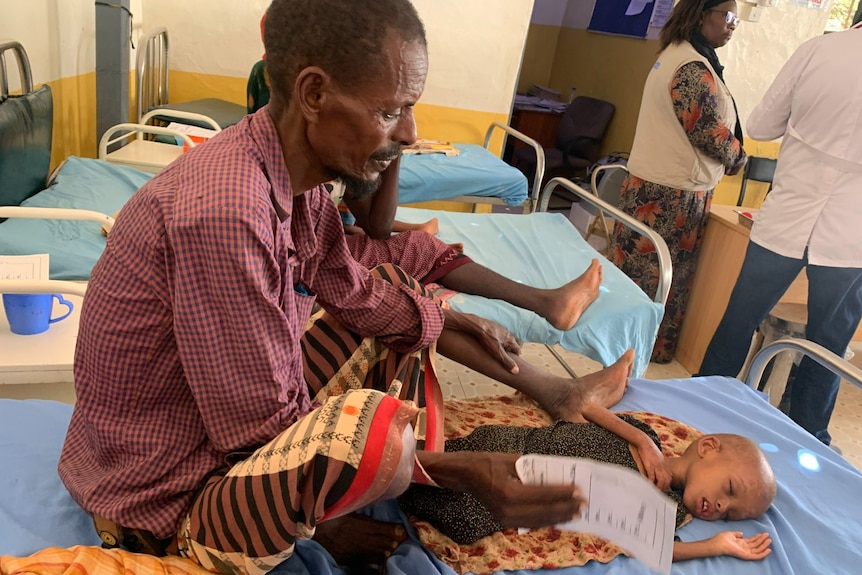 Man sitting on a hospital bed with his very sick daughter lying asleep in front of him.