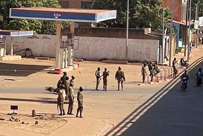 Men in military uniforms stand in the street near a petrol station. 