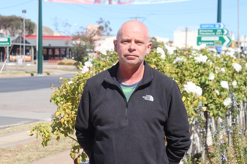 A bald man wearing a jumper standing in front of a flower garden and street